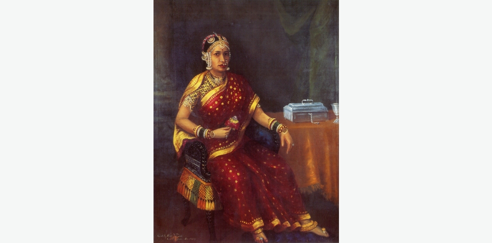 A portrait of an ornately dressed woman in a red saree sits in an armchair and looks at the viewer while holding a flower in her right hand. There’s a table with a jewellery box and a goblet on her left.