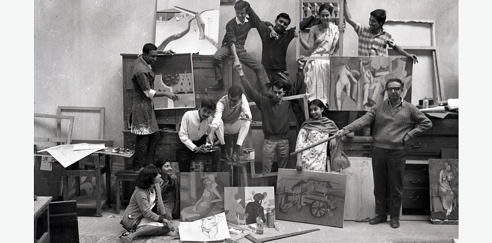 A group of students sitting on the floor pose for the camera. The shelves in the room showcase their paintings.