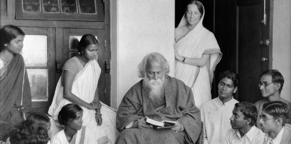 Seated on a chair, a white-bearded man in a loose robe reads a book surrounded by students.
