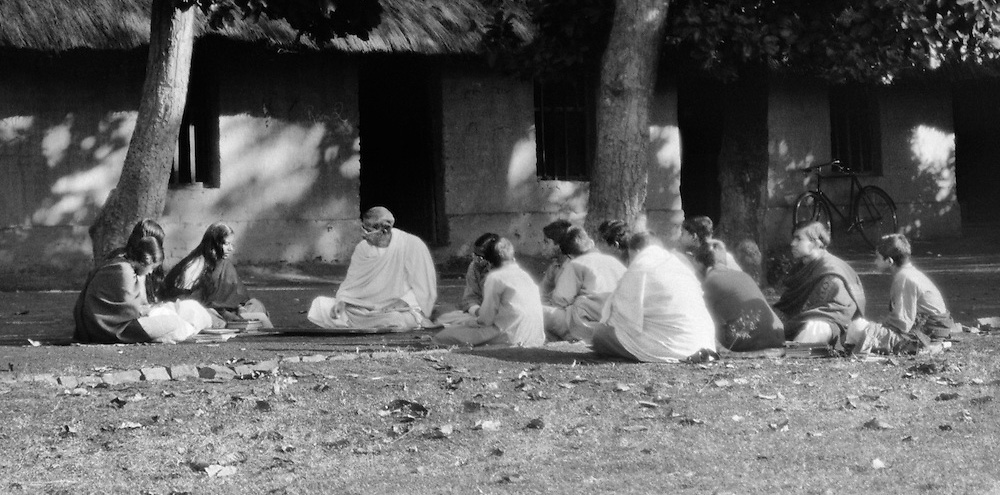 A group of students sit on the ground in discussion with their teacher surrounded by a thatched hut and trees.