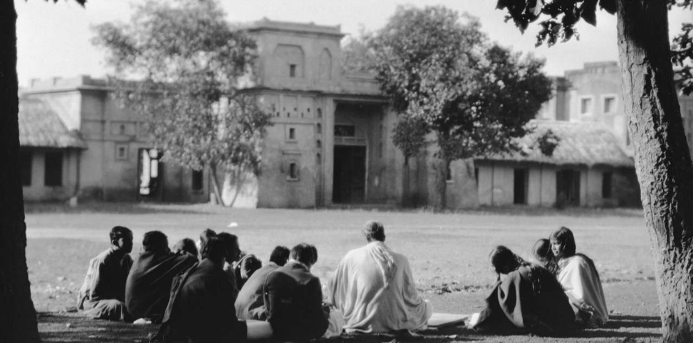 A group of students and their teacher with their backs facing us sit in the shade of trees outside a building.