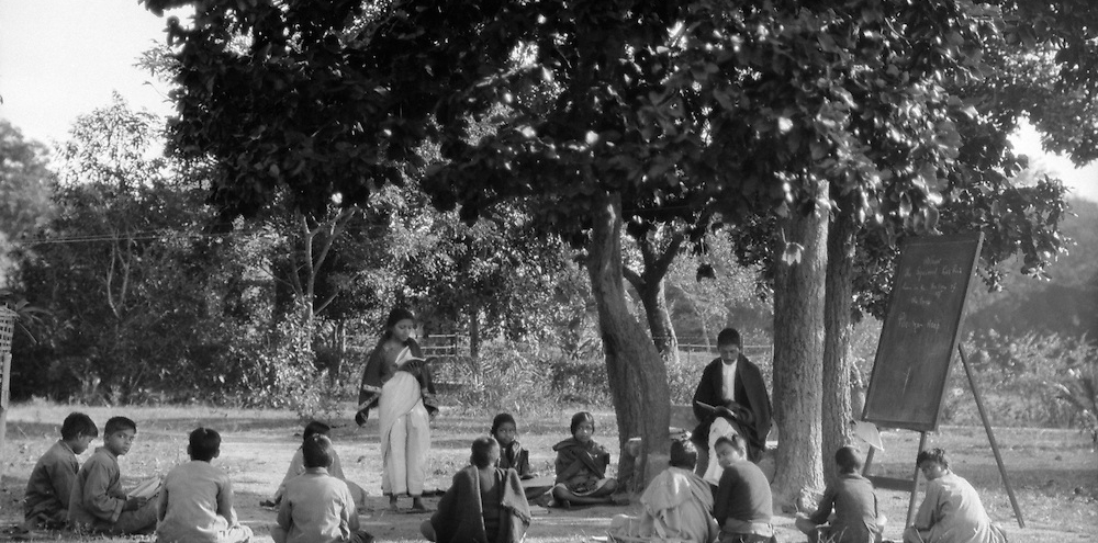 A group of seated students surround their teacher and a blackboard in the shade of trees while a girl stands and narrates from a book.