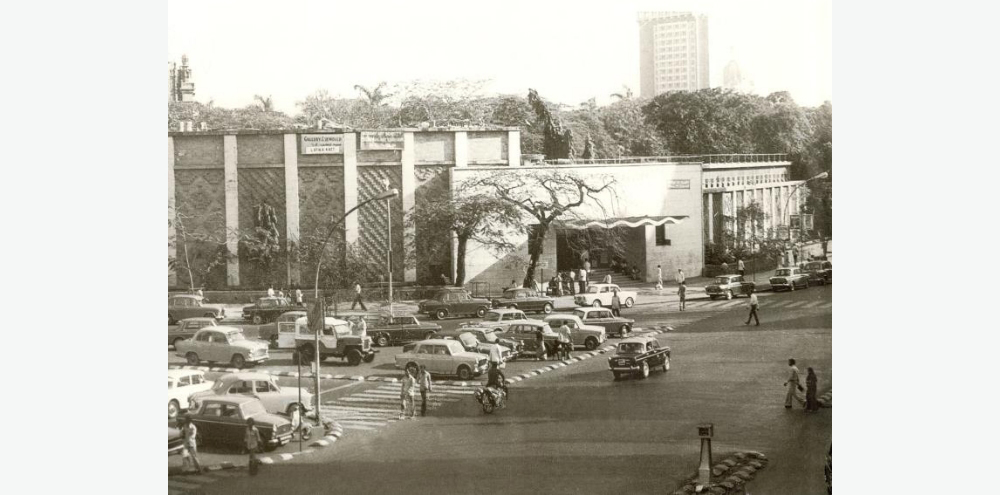 Multiple cars stand in a parking lot in front of a large building, with a scattered group of people walking and crossing roads.