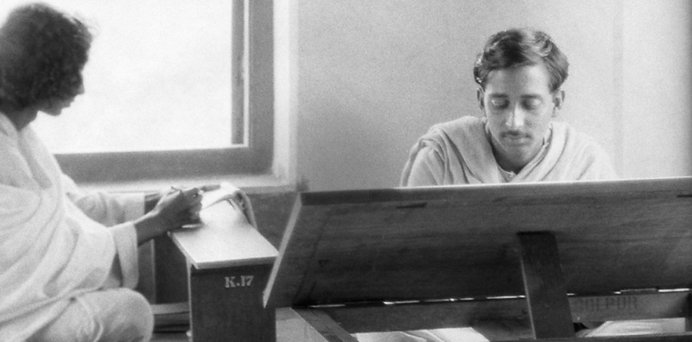 Two male art students dressed in white sit on wooden benches and work in a room with a large window in the left background.