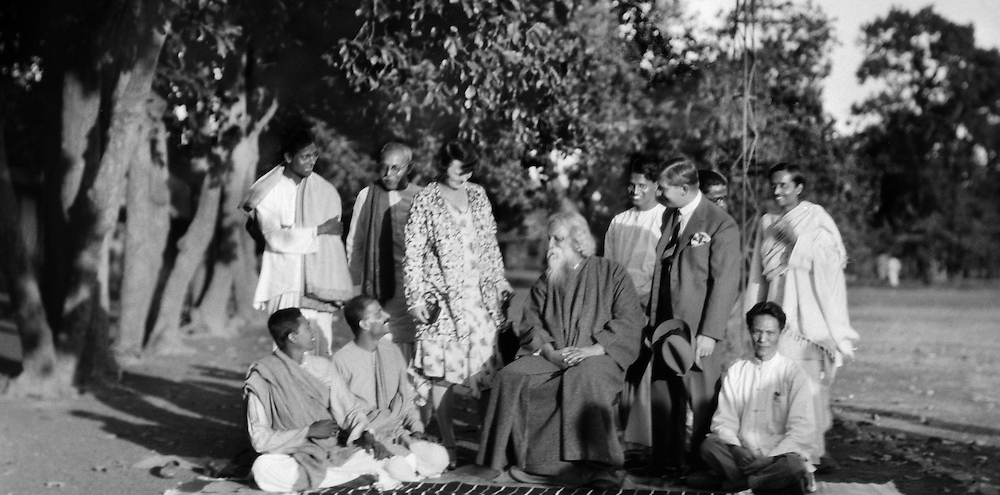 A bearded man sits on a chair in the centre, surrounded by Indian and international professors with a row of trees on the left.