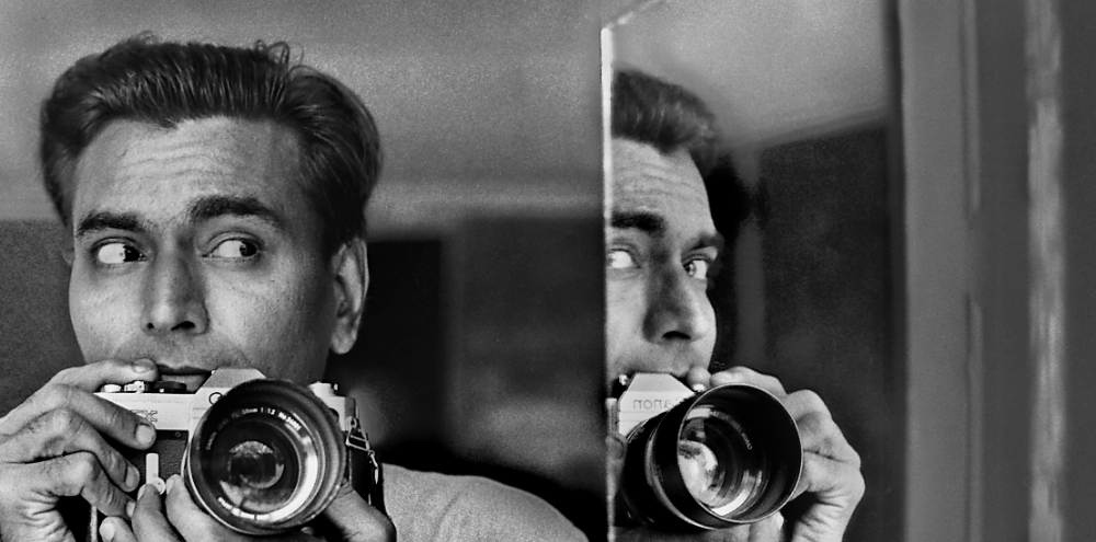 The artist adjusts his camera lens and presses the shutter button while looking left at his reflection in a mirror angled towards him.