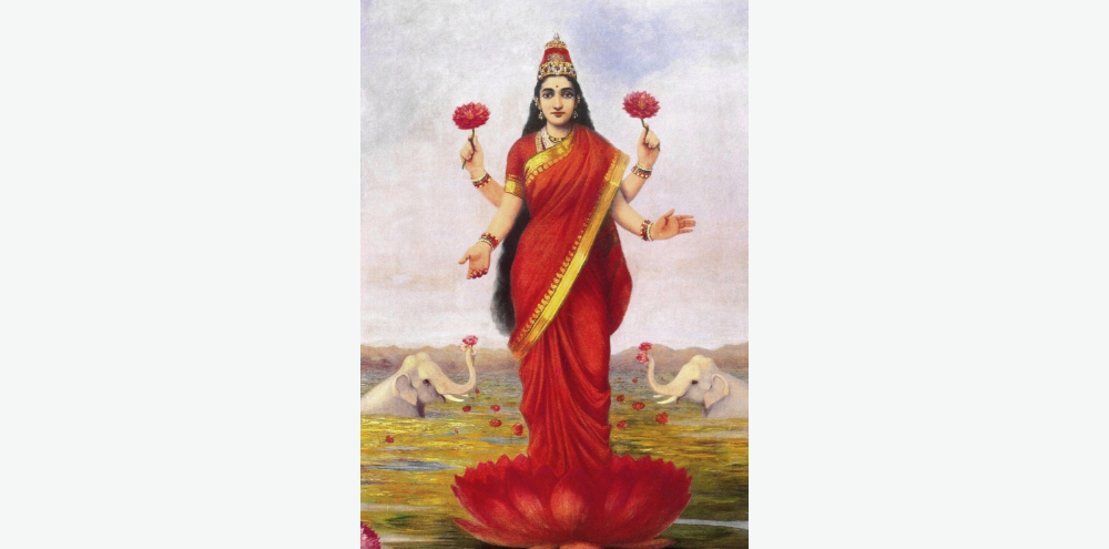 A four-armed woman dressed in a red saree holds lotuses in two of her hands while standing on a large lotus in a pond. She is flanked by two half-submerged white elephants raising lotuses in their trunks in the distance.