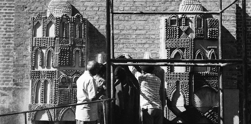 A group of men assemble panels of a relief mural on the brick facade of a building with scaffolding in the foreground.