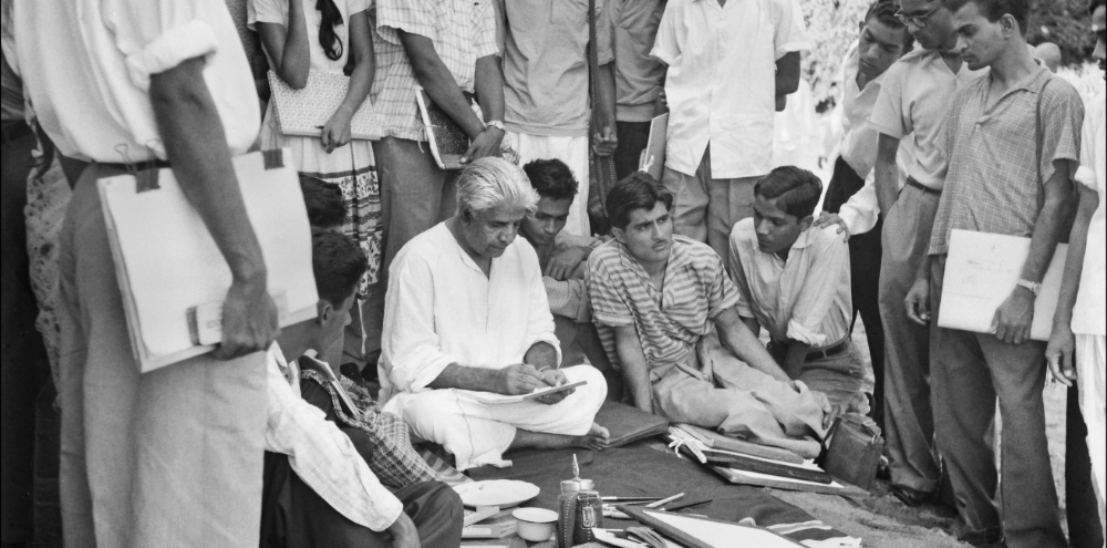 A group of students with their sketchbooks surround and observe a seated man in white garments drawing on paper.