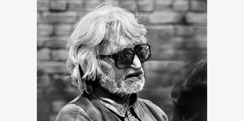 A man with grey hair and a beard wearing black sunglasses looks beyond the frame towards the right.