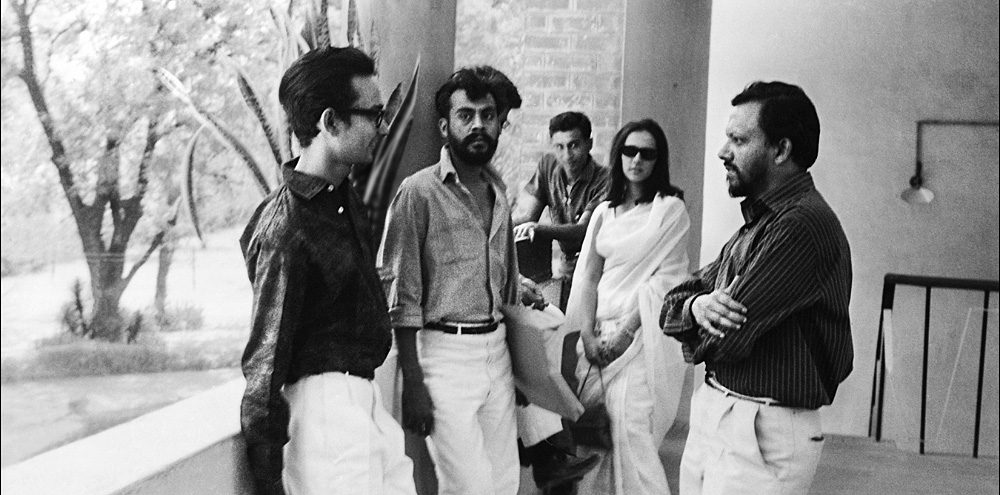 A group of men and a woman lean against the parapet of an open corridor and engage in conversation facing each other.