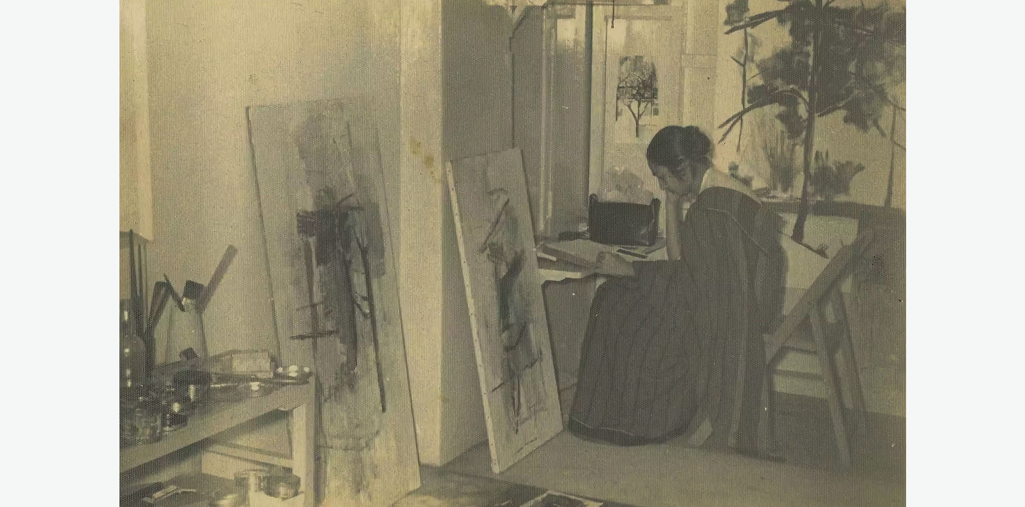 A woman sits in contemplation on a chair at her desk in her studio full of abstract paintings and a table with art supplies.