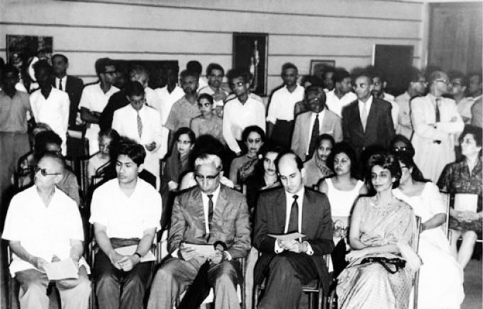 A large group of seated and standing people formally dressed as they look at something beyond the frame to our left.