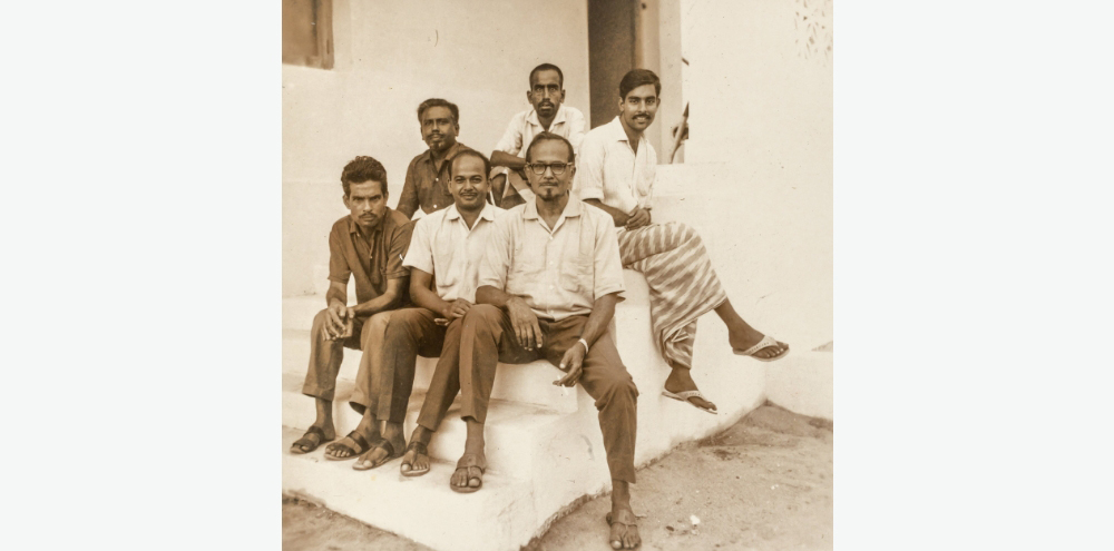 A group of six men sit on a staircase outside a building, and face towards the camera.