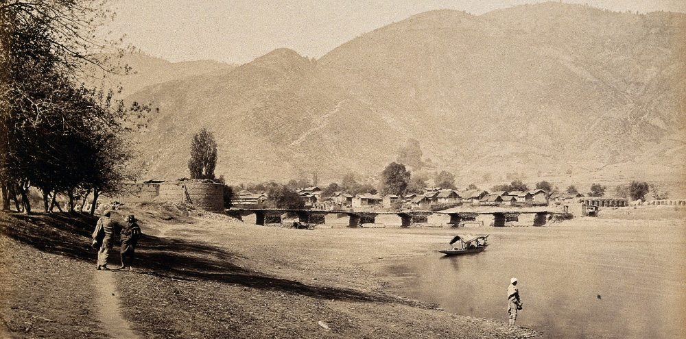 On the left, two people walk on the shore of a river while a boat, bridge and row of houses are on the right with large mountains in the background.