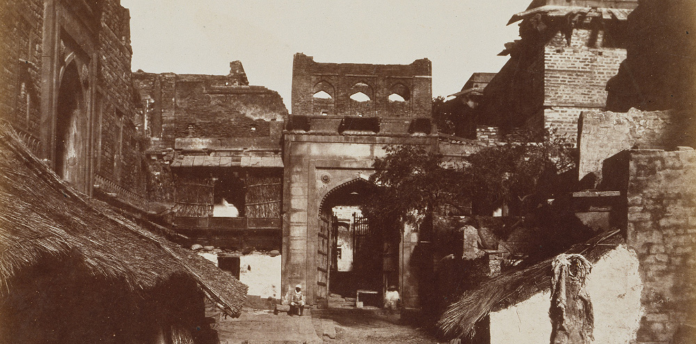 A sepia toned photograph of old multi story brick buildings with thatched roofs, and a large gateway in the centre of the buildings.