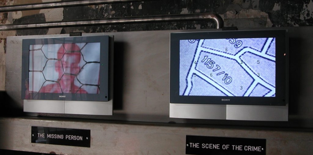 Two wall screens with stills showing a man behind a grill and a grid with numbers, labelled with the English text, ‘Missing Person’ and ‘The Scene of the Crime’ respectively.