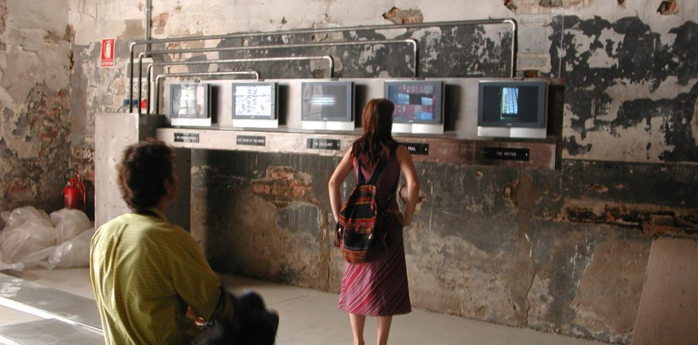 A seated man and a standing woman observe five labelled screens connected to pipes installed on a plaster-chipped wall with an electric box on the far left.