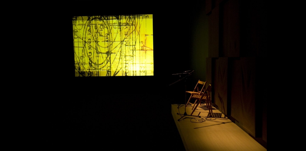 A wooden chair is spotlighted in a dark room with a video projection of superimposed black text and shapes on a bright yellow background.