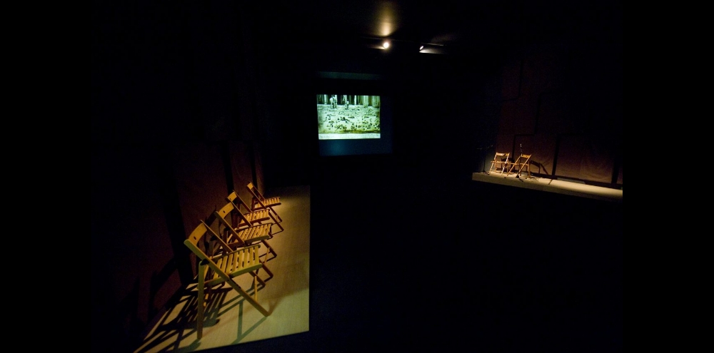A dark room with spotlit wooden chairs converging towards a screen projection in the background.