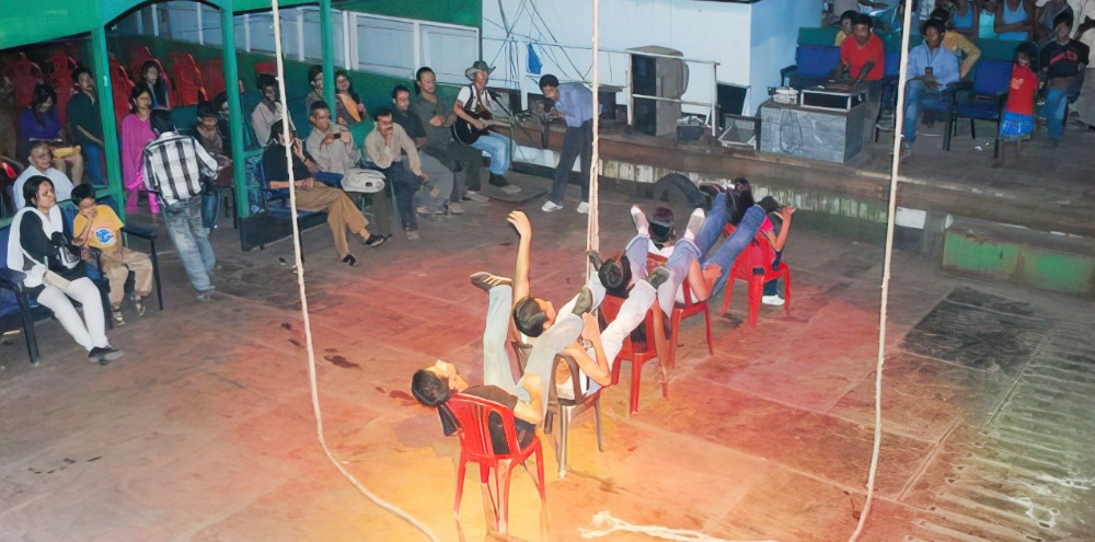 Five people sitting on red chairs raise their legs in the air while a large group of people on the left watch them.