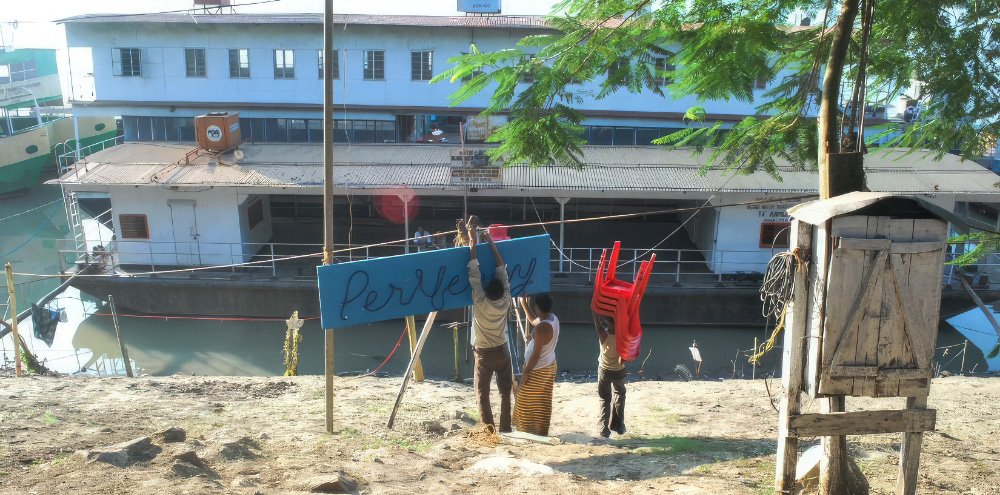 Two people hang up a sign with the English text, ‘Perriferry’ in front of a tin roof white boat with water around it.