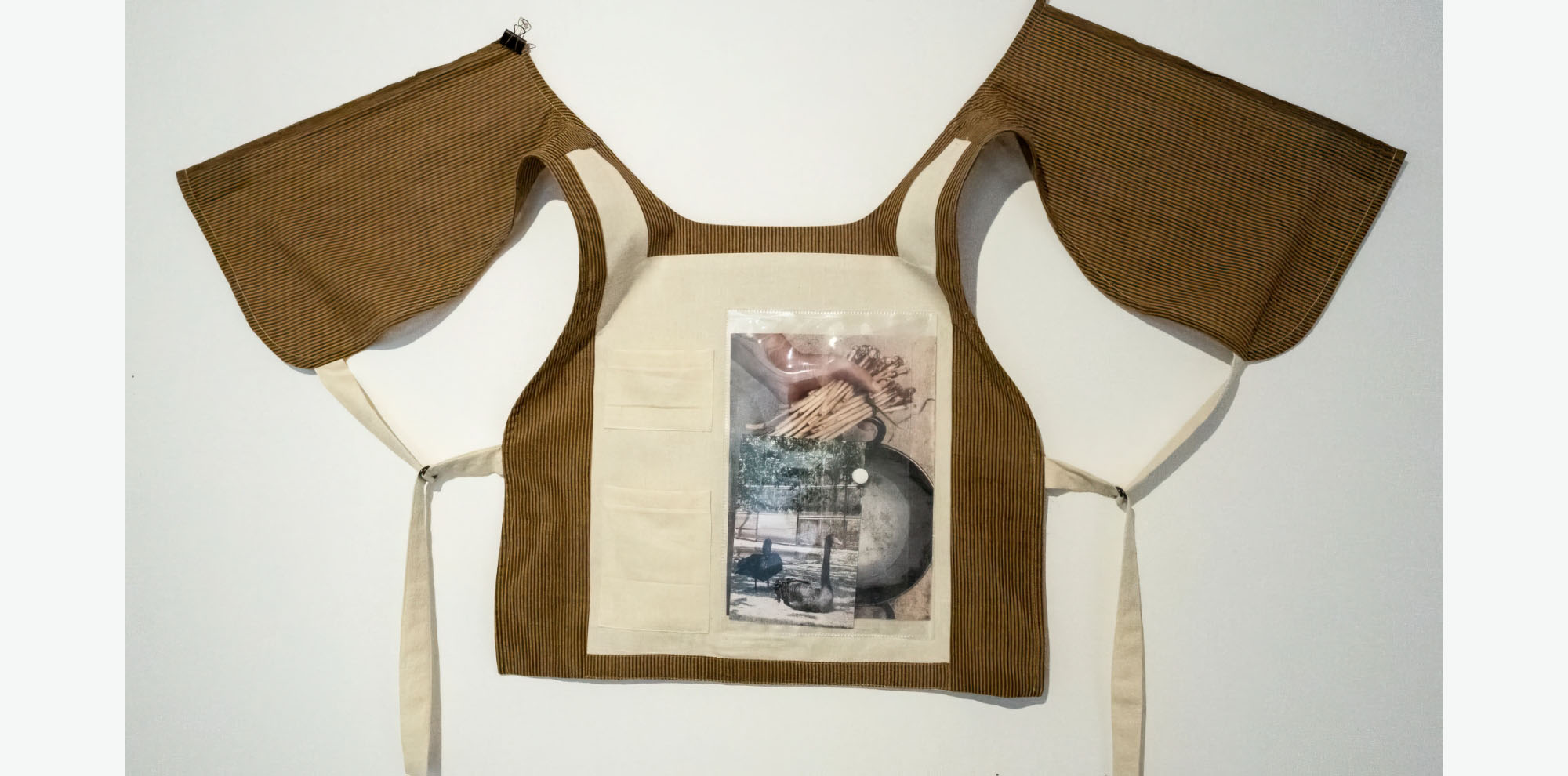 A vest with a transparent pocket hangs against a white wall. The pocket contains a photograph which depicts a hand holding sticks over a vessel, as well as a small photograph of two birds.