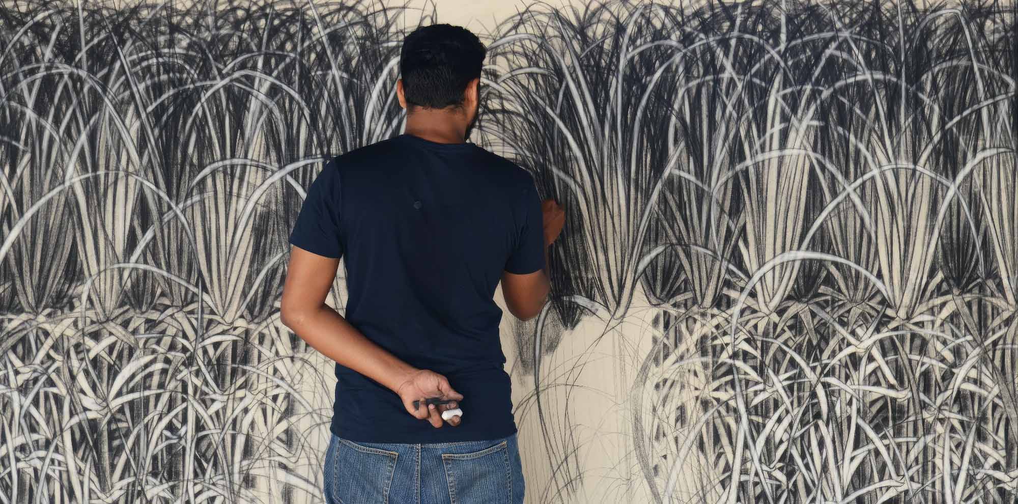 A man draws grass with charcoal and chalk on a wall with his back facing the viewer.