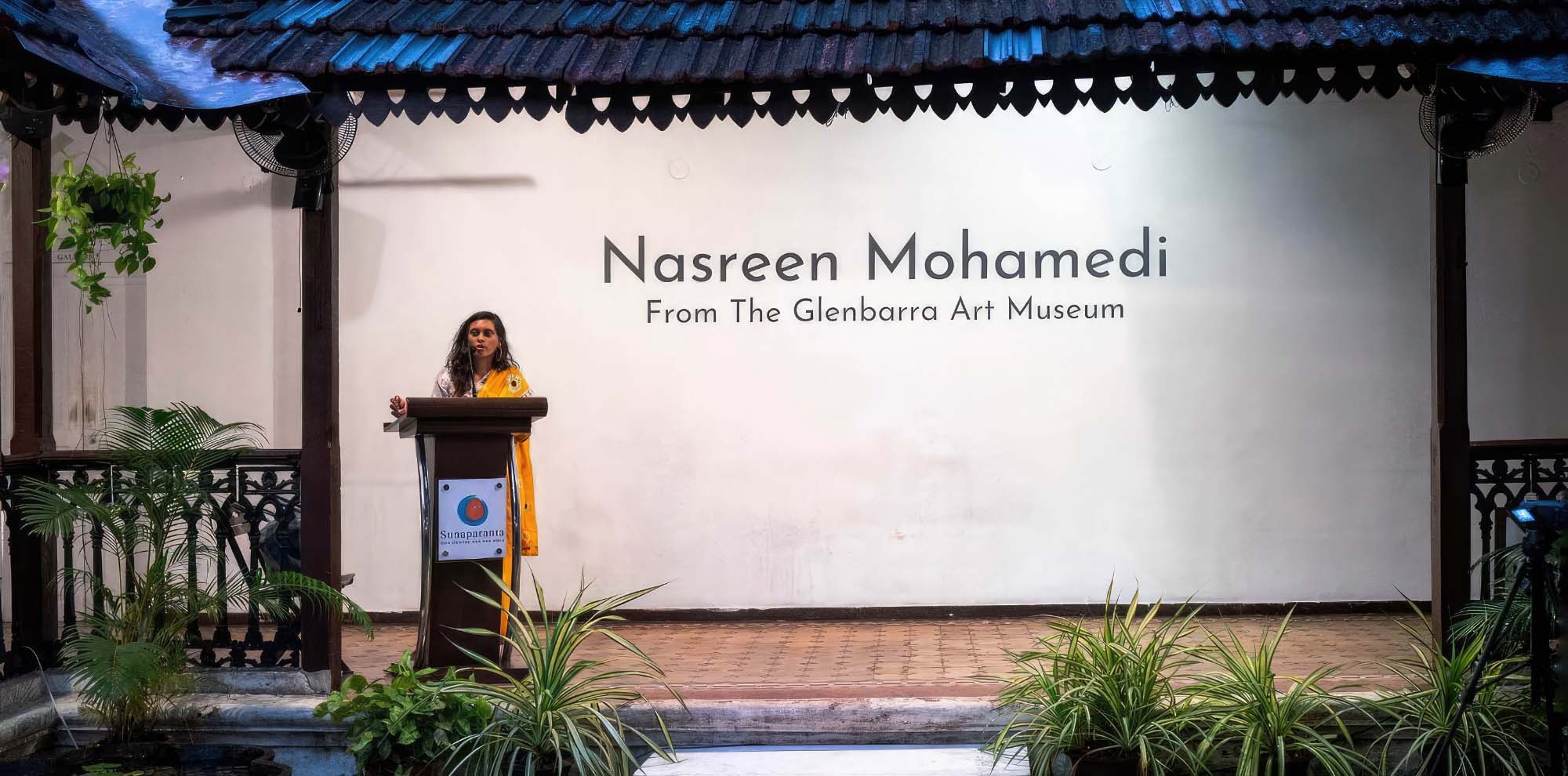 A woman gives a speech on a podium in an open corridor with the English text ‘Nasreen Mohemadi: From the Glenbarra Art Museum’ on the wall behind her.