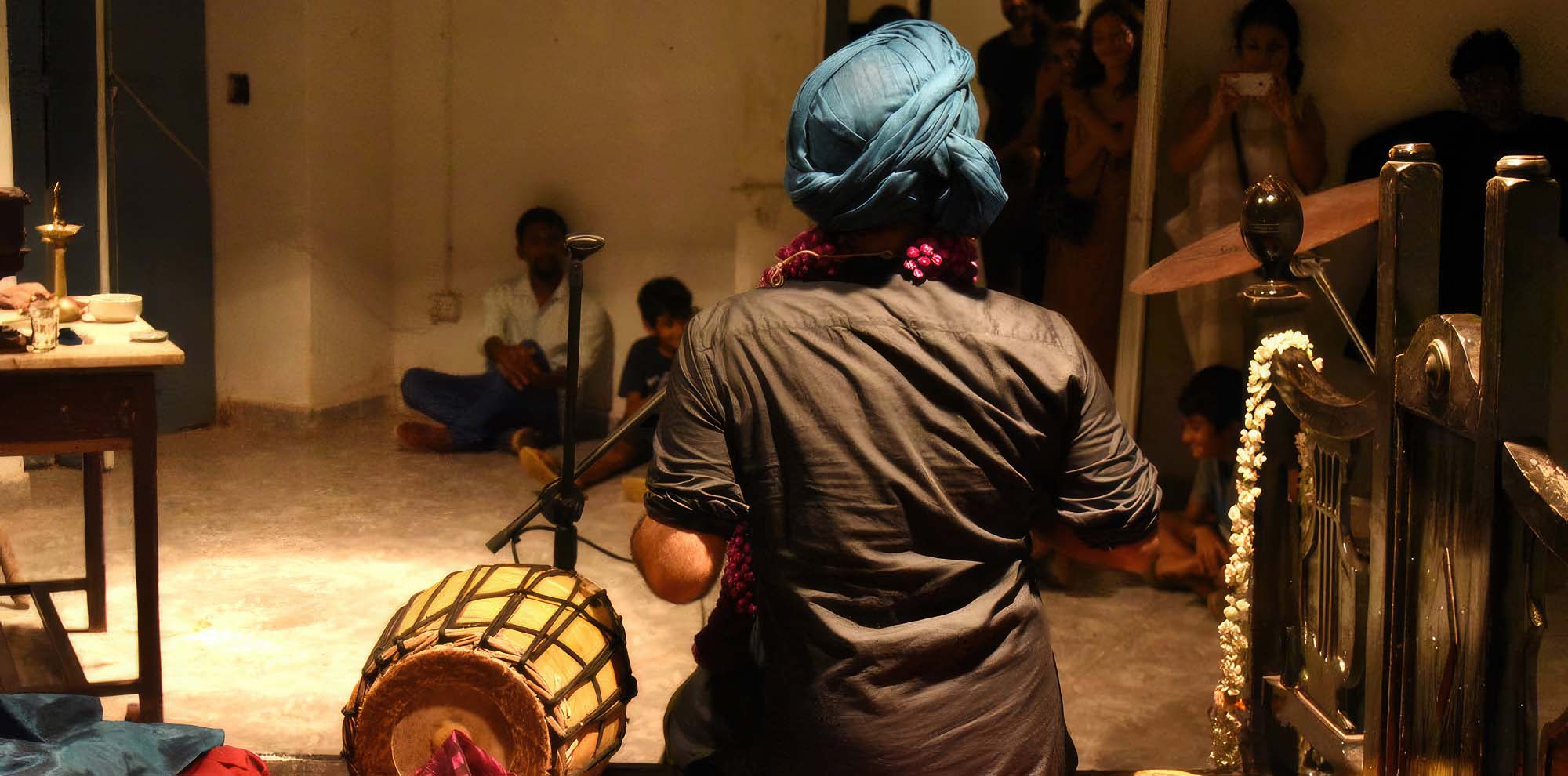 The artist sits on a bed with his back to the viewer, a dhol to his left and a group of people watching him perform at the other end of the room.