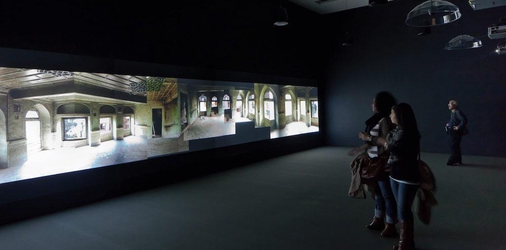 Three people in a dark room watch a video projection of different interior sections of an abandoned building.