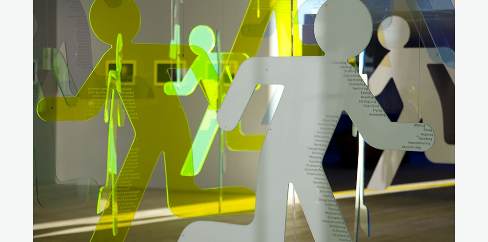 White and yellow cutouts of running figures with printed words towards the edge hang in an exhibition space.