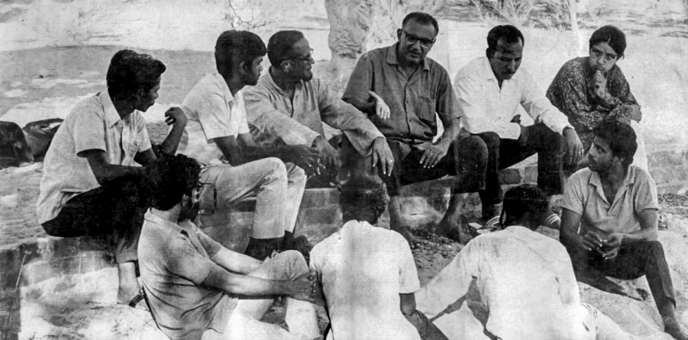 A group of men are seated as they engage in deep conversation under the shade of trees.