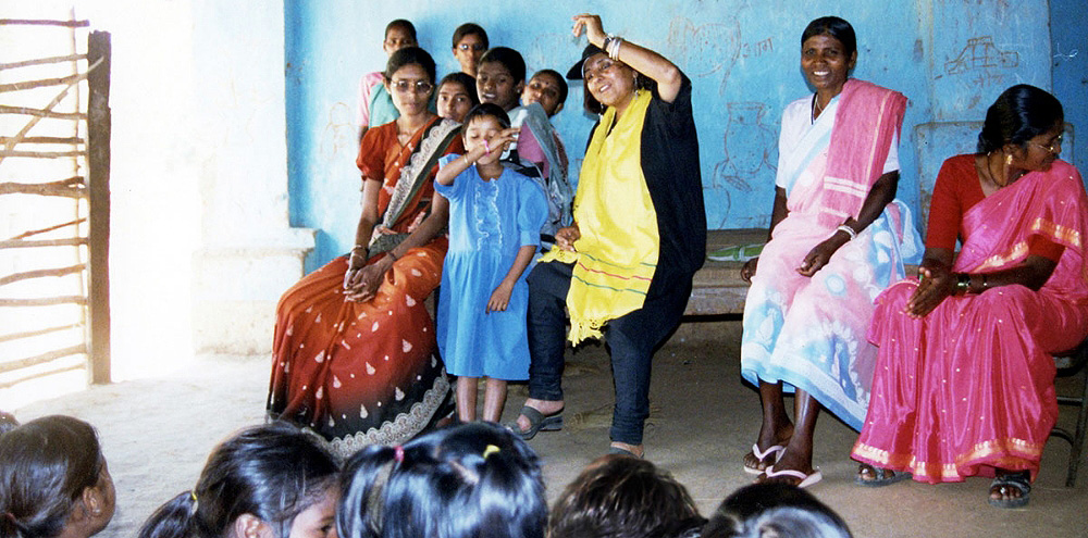 A group of women and children in a classroom listening to a girl recite.