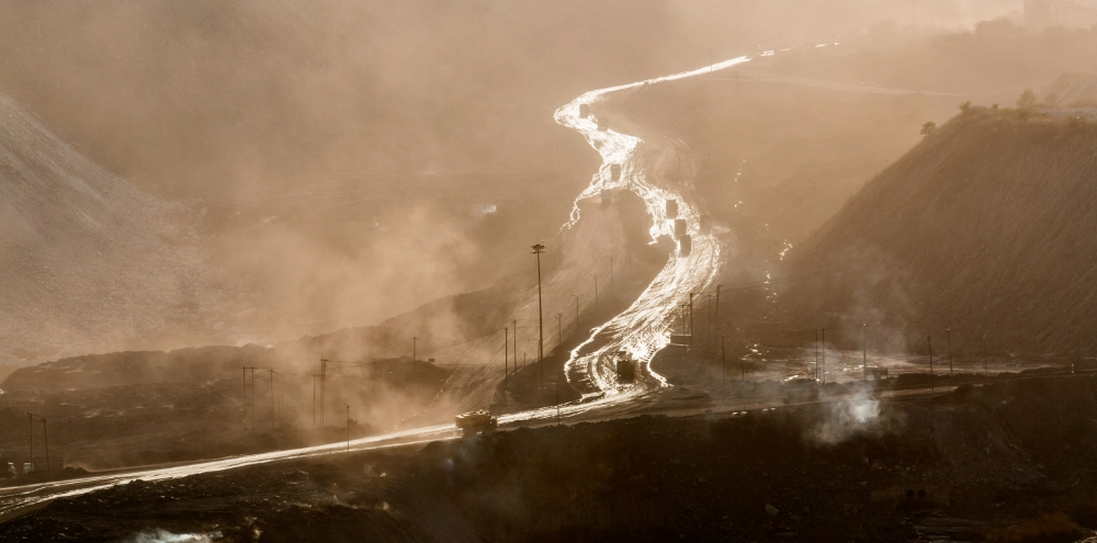 A landscape shot of mountains with winding roads, and trucks carrying mined material. The road seemingly glows under the sun which is shrouded by smoke clouds.