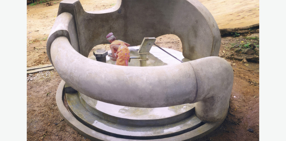 A pipe-shaped concrete circular wall surrounds a hand-operated water pump where a woman collects water.
