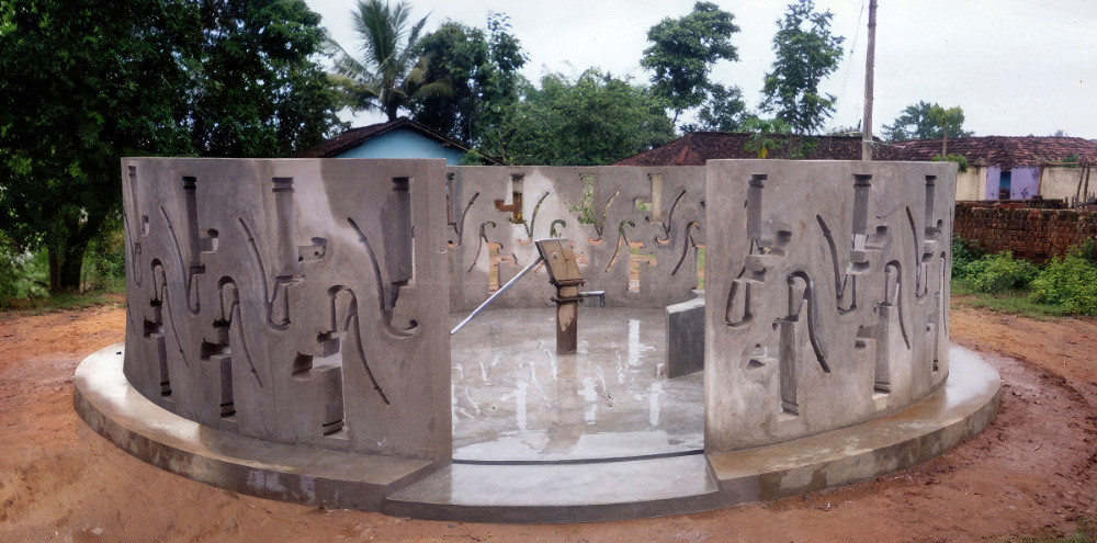 A circular concrete wall with water pump-shaped holes surrounds a functioning hand-operated water pump.