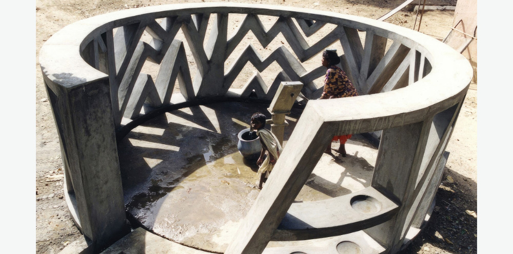 A circular wall with irregular chevron-striped openings surrounds a hand-operated water pump where a woman and child are collecting water.