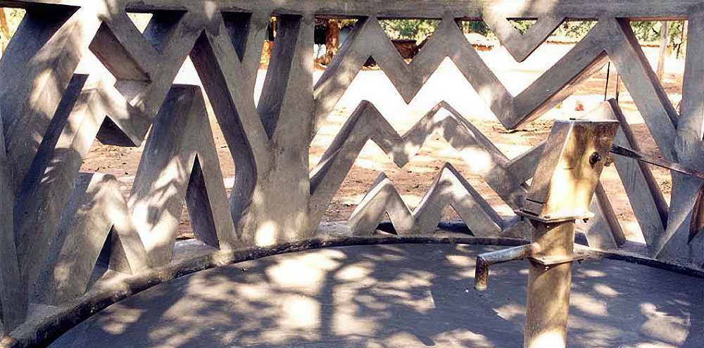 A hand-operated water pump with a circular wall with irregular chevron-striped openings in the background.