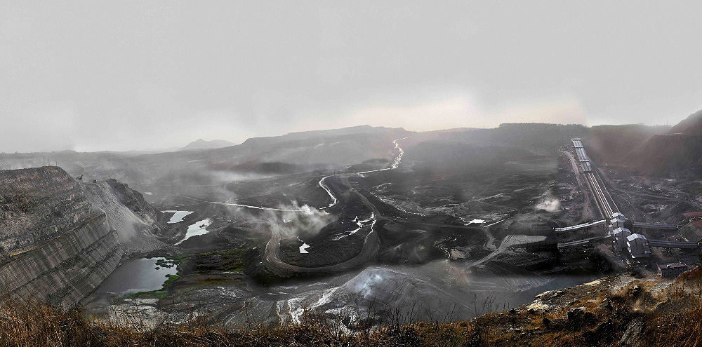 A landscape shot of large black mountains with winding roads. The mountains are being mined and there’s a train station to the far right.