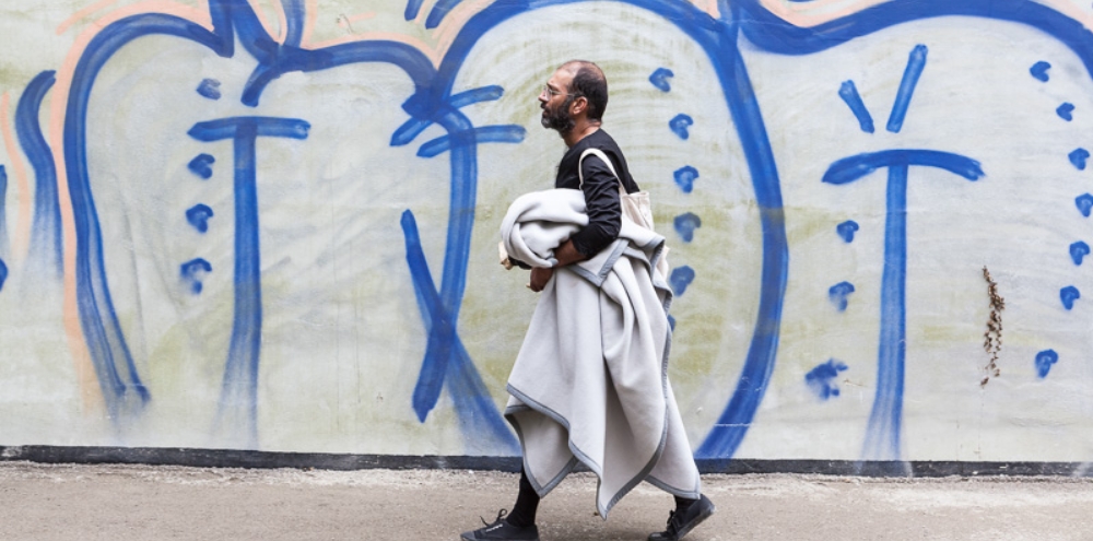 A man carrying a blanket and bag walks in front of an abstract mural.