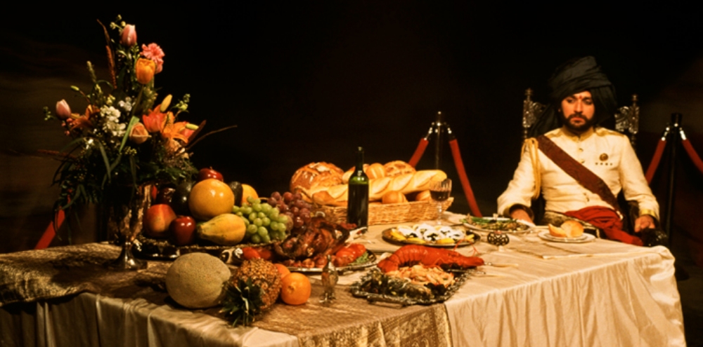 A seated man dressed in royal garments and a turban looks at a table full of fruits, lobster, bread and wine.