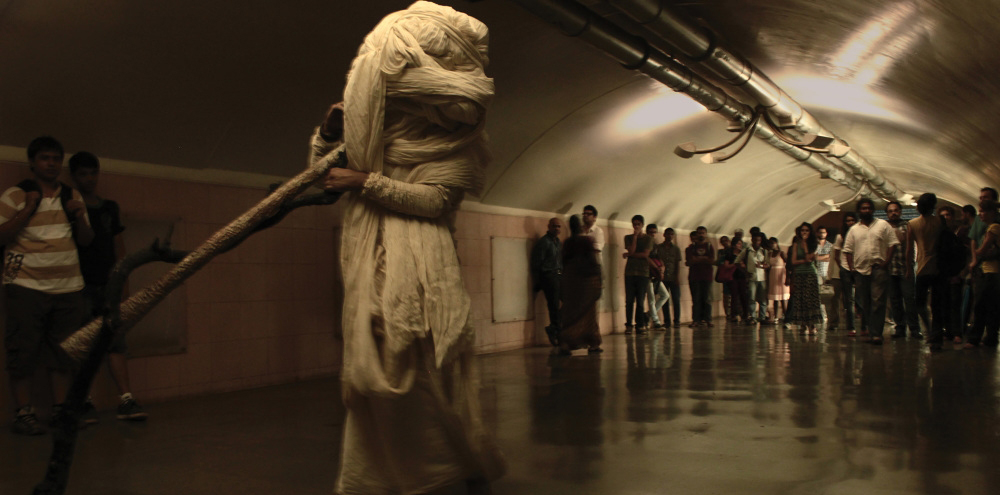 A man wrapped entirely in white cloth walks through a tunnel with an audience observing him in the background.