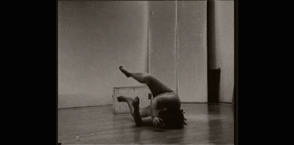 A nude woman with her legs raised and arms bent falls on the floor on her side.