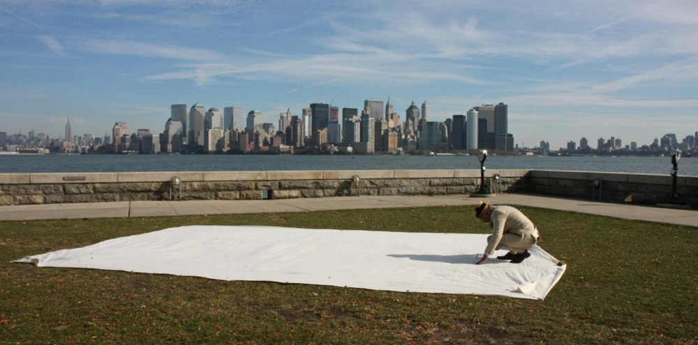 A crouching man draws on a large cloth with a river and high-rise buildings in the background.