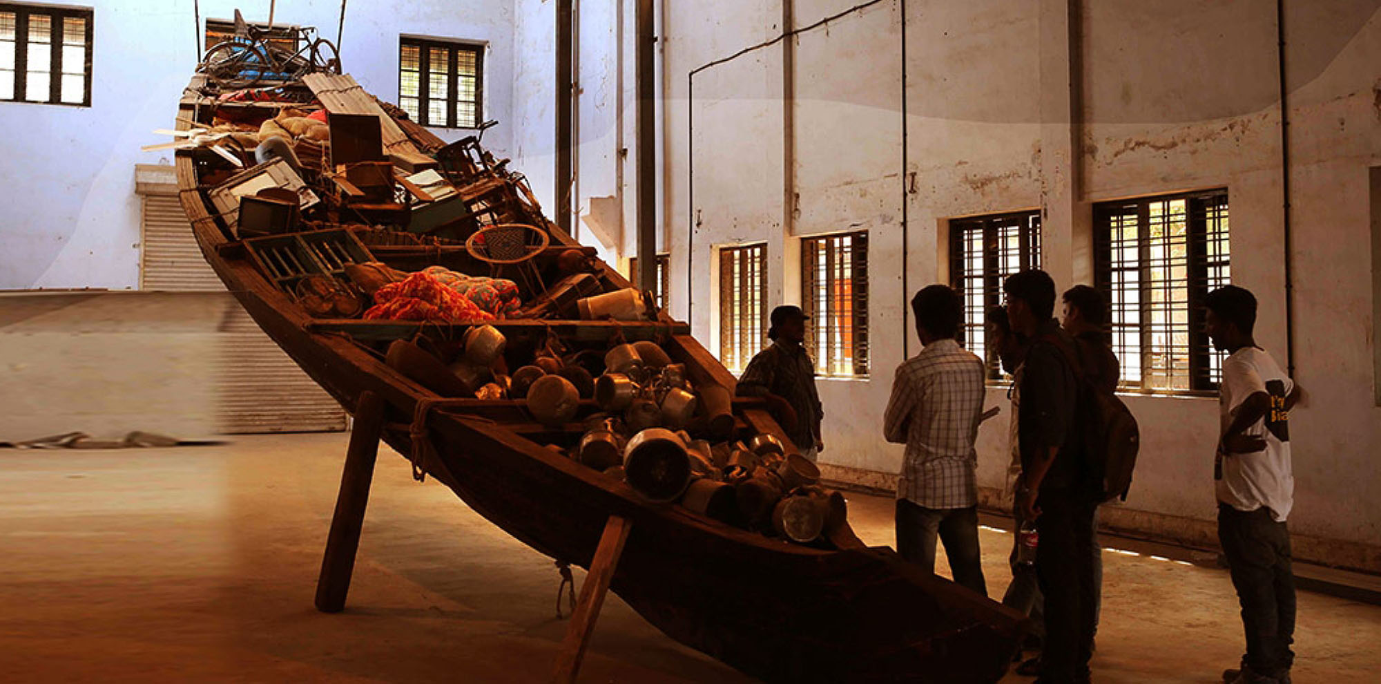 A boat filled with various objects was lifted higher and placed in a large room with a group of men chatting beside it.