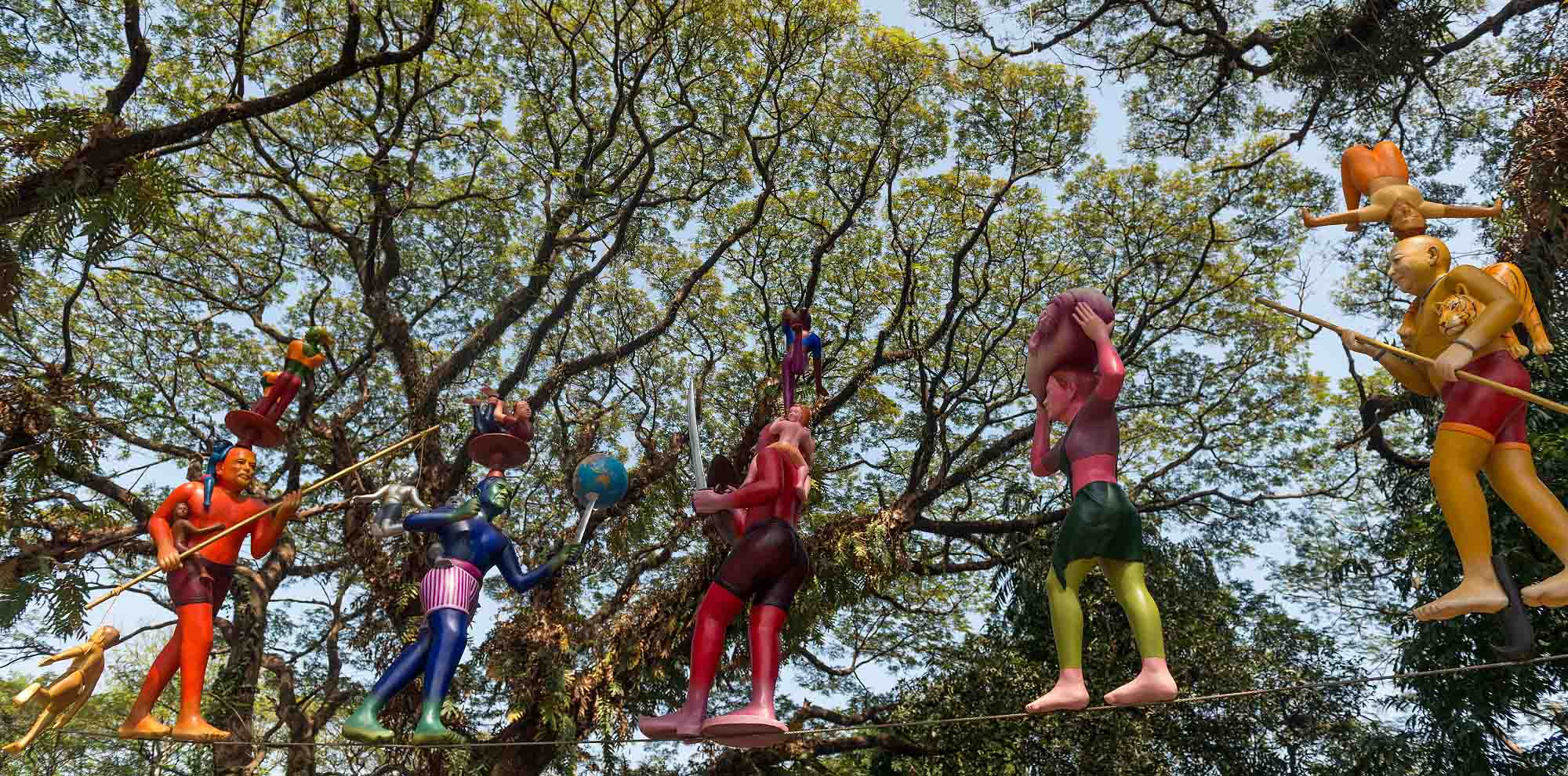 Brightly painted human-like sculptures balance various objects on their heads and walk across a tightrope with trees in the background.