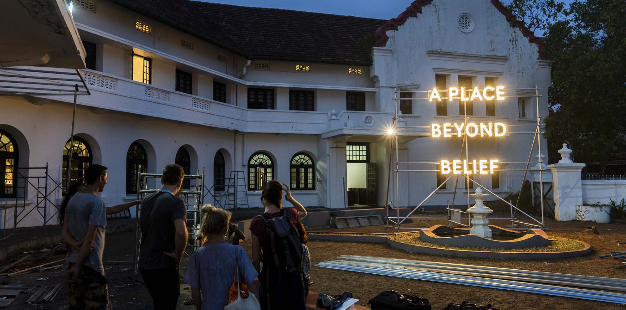 An illuminated sign with the English text, ‘A Place Beyond Relief’ is installed on a metal framework outside a white building with a group of people admiring it.