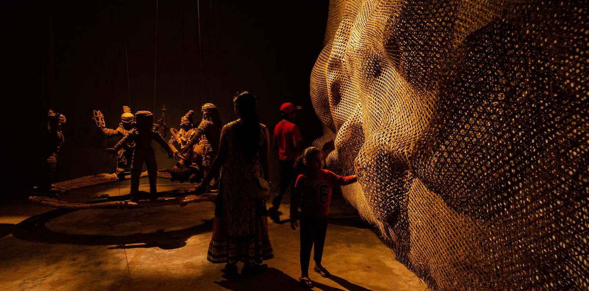 People admire a bulbous wall on the right with puppet-like sculptures made from bamboo, jute and straw standing in a circle in the background.