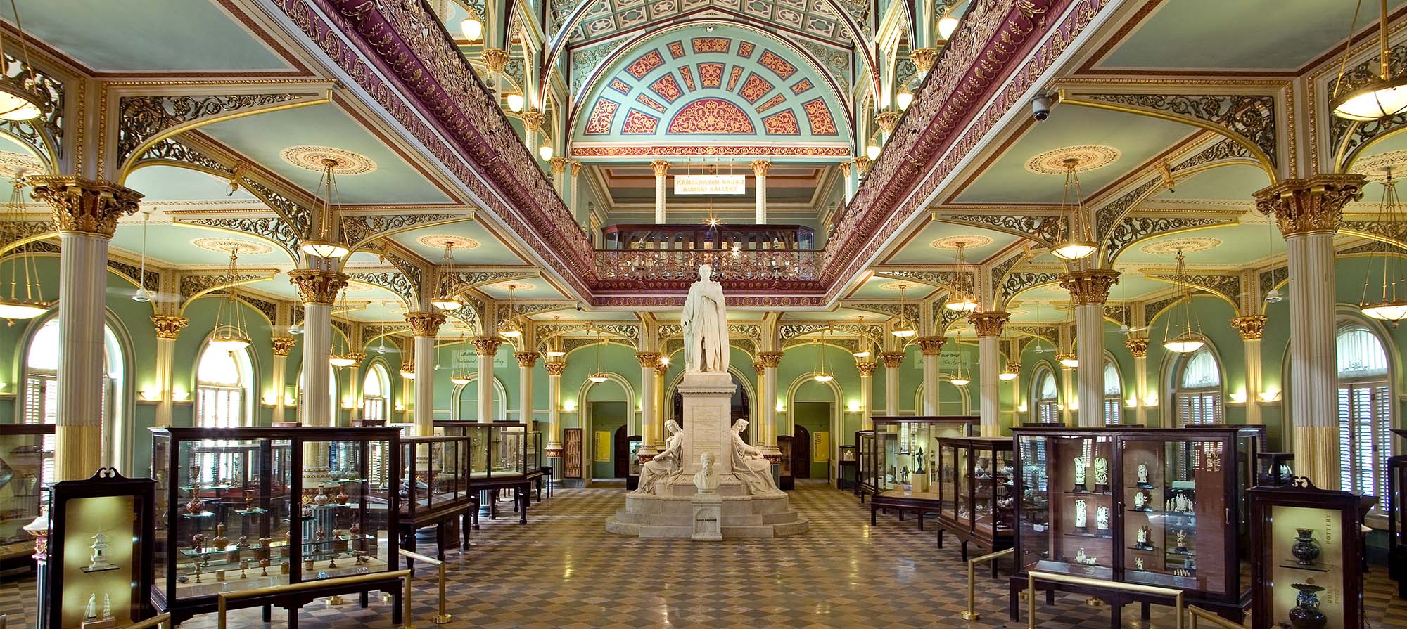 A long hall with brightly painted blue walls, decorative ceilings, gilded columns and a large statue of a man surrounded by glass cabinet displays.